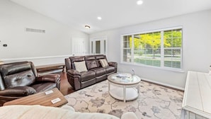 A spacious living room with a brown leather sofa, a glass coffee table, and a large window with blinds. - Cedar Falls Retreat: Beachfront 5BR Home w/ Community Beach Access (Post Falls)