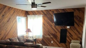 A living room with a wooden wall, a ceiling fan, a flat-screen TV mounted on the wall, a sofa, a chair, and a window with curtains. - Home on Yucca (El Centro)