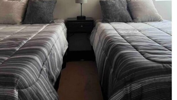 A bedroom with two beds, a nightstand, a lamp, and a painting of a sunset over a pier. - Home on Yucca (El Centro)