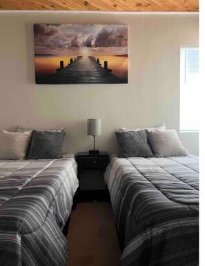 A bedroom with two beds, a nightstand, a lamp, and a painting of a sunset over a pier. - Home on Yucca (El Centro)