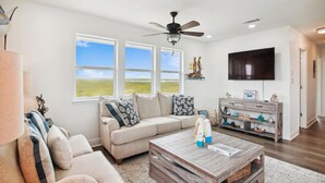 A spacious living room with a large window, a ceiling fan, a flat-screen TV, and a wooden coffee table. - Blue Fin by Ryson Vacation Rentals (Freeport)