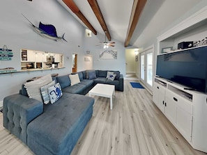 Uma sala de estar moderna com um sofá modular, uma TV de tela plana, vigas de madeira no teto e uma cozinha ao fundo.