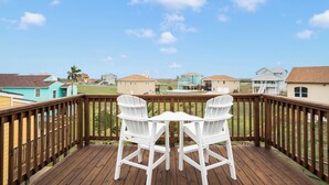 A wooden deck with two white chairs and a table, overlooking a residential area with houses and a clear sky. - Sea Breeze Escape by Silver Sands Vacation Rentals (Port Aransas)