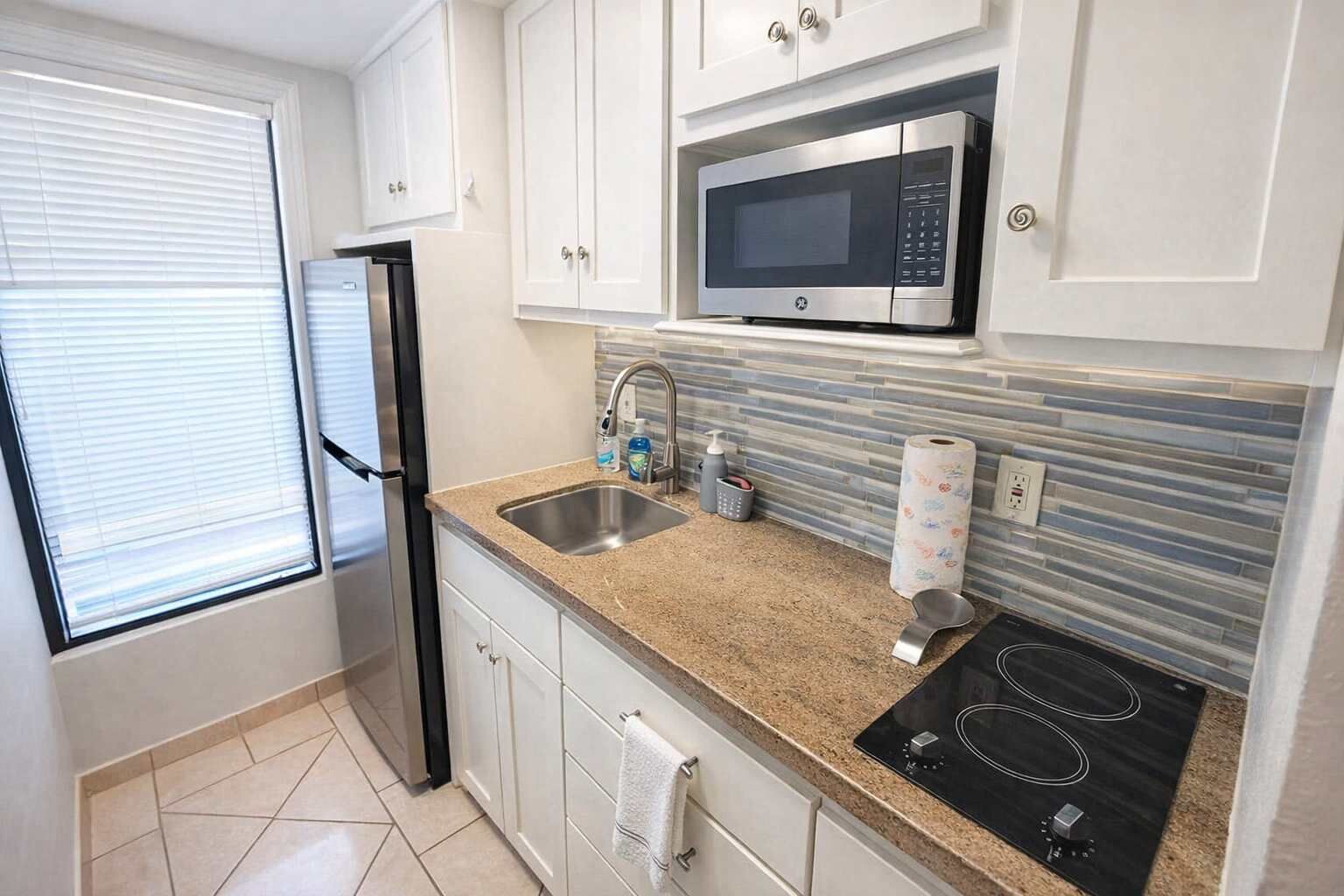 A compact kitchen with a stainless steel refrigerator, a microwave, a sink with a faucet, a countertop with a soap dispenser, and a tiled backsplash.