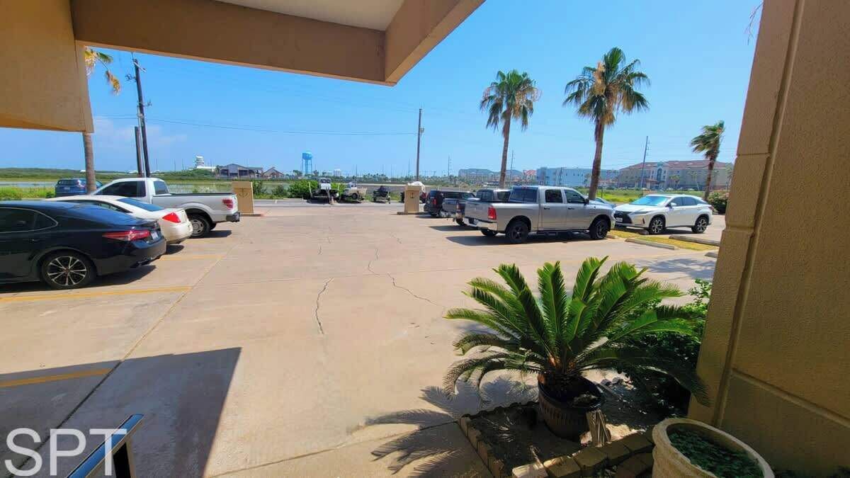 Ein Parkplatz mit mehreren Autos, darunter eine schwarze Limousine und ein weißer Pritschenwagen. Im Hintergrund sind Palmen und ein Gebäude zu sehen.