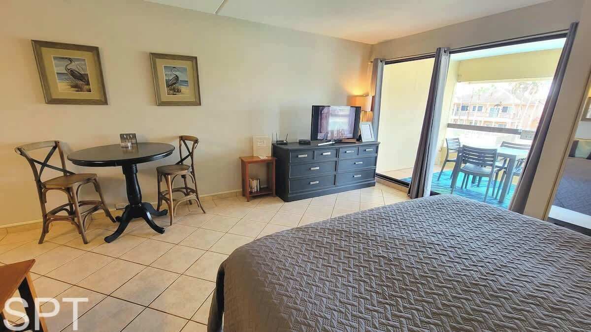 A hotel room with a bed, a desk with a chair, a television, a dresser, and a balcony with chairs.