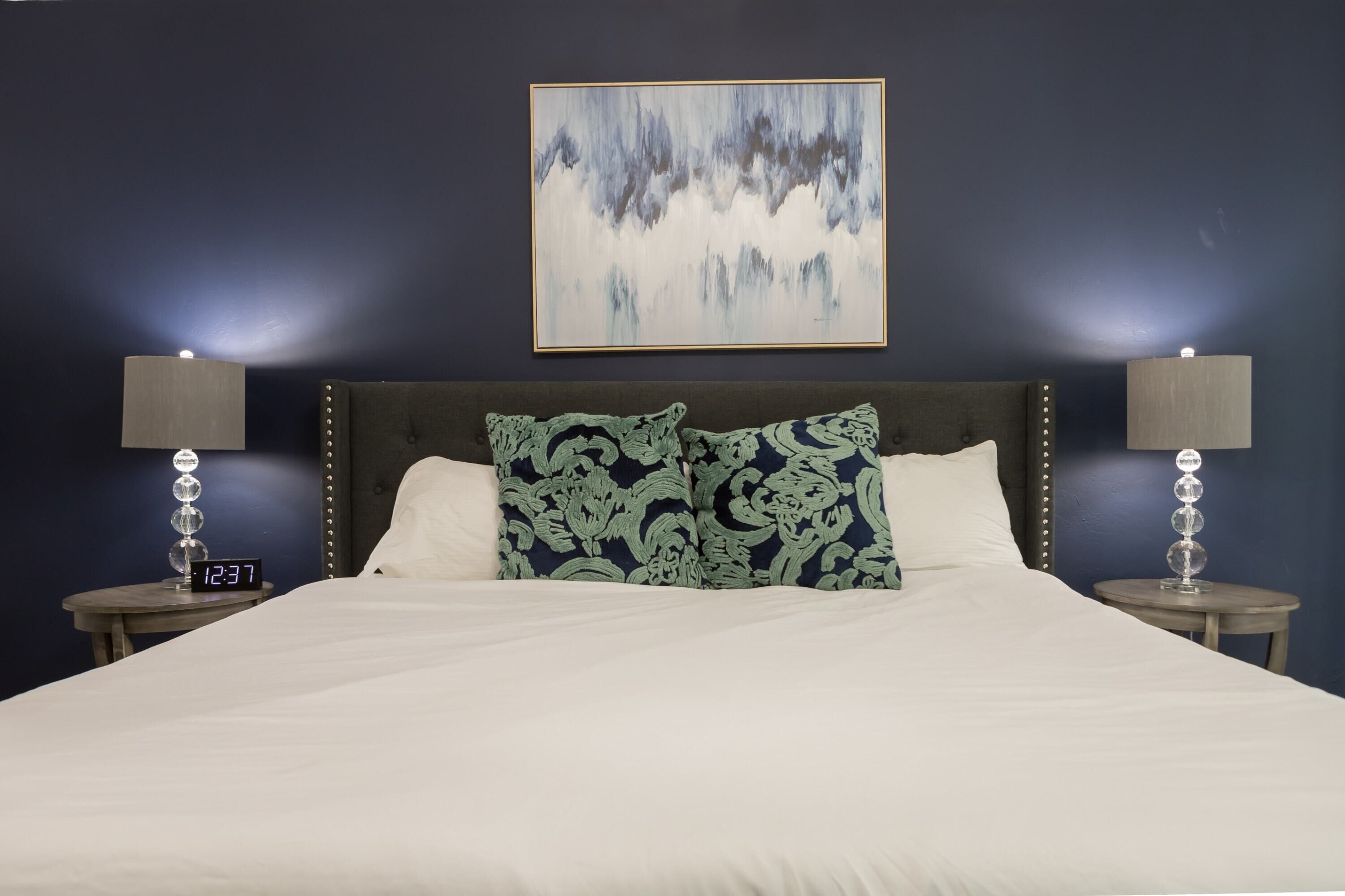 Una cama bien tendida con sábanas blancas y dos almohadones decorativos, flanqueada por dos mesitas de noche con lámparas y relojes, frente a una pared azul oscuro con un cuadro abstracto enmarcado.