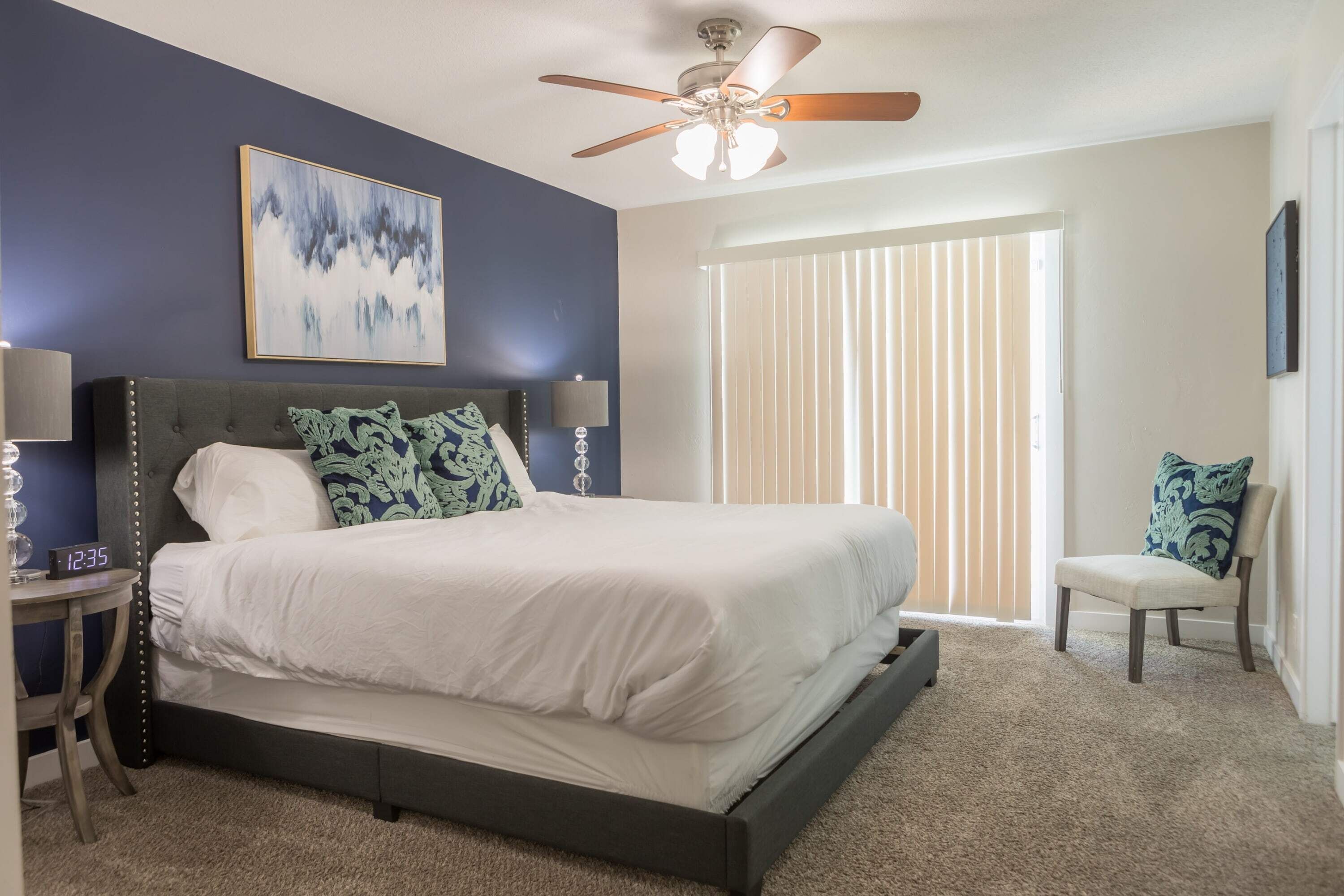 Une chambre à coucher comprenant un lit, un ventilateur de plafond, une table de chevet, une chaise et une fenêtre avec des stores.