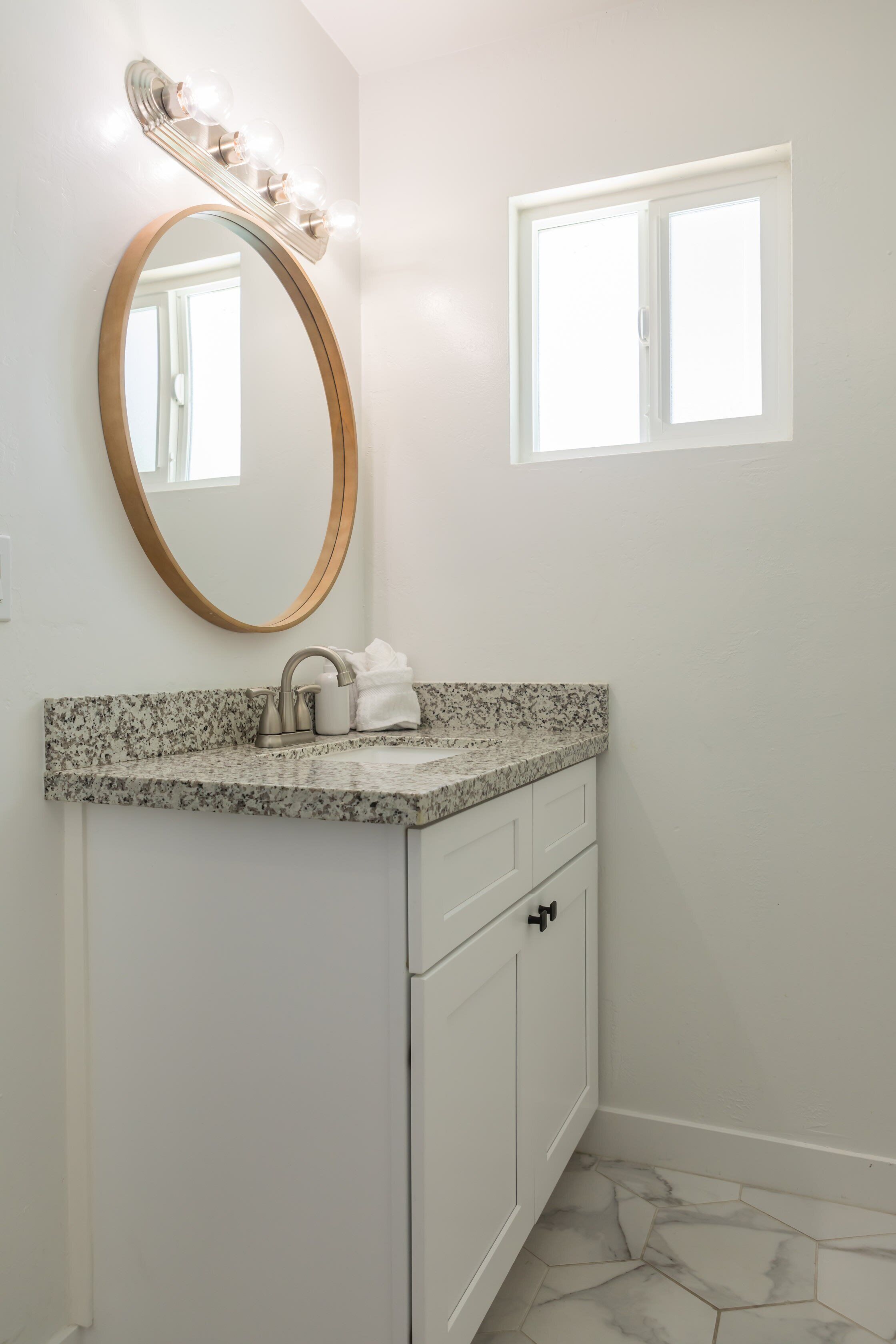Une salle de bain avec un comptoir en granit, un grand miroir rond avec éclairage intégré, un lavabo blanc et une fenêtre.