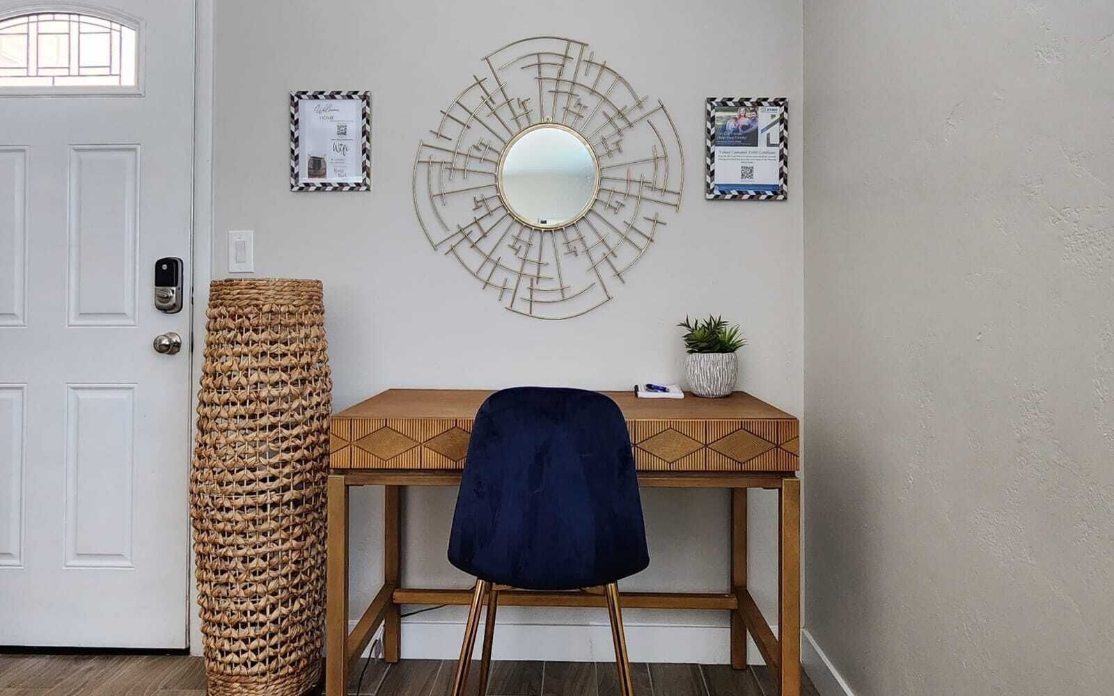 Une pièce avec un bureau en bois, une chaise bleue, un panier en osier, un miroir fixé au mur et deux certificats encadrés accrochés au mur.