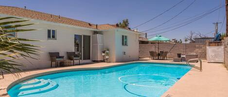 A swimming pool with a curved edge, a patio area with chairs, and a building with a light-colored exterior.
