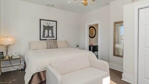 Ein Schlafzimmer mit einem Bett, einem Nachttisch mit Lampe, einem Sessel, einem Spiegel und einem gerahmten Bild an der Wand.