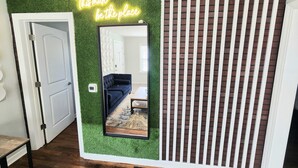 Quarto com parede verde, letreiro neon, espelho, sofá azul escuro e divisória com padrão de tijolos.