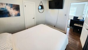 Quarto com uma cama grande, televisão de tela plana na parede, uma porta e uma pequena escrivaninha com luminária.