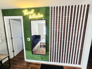 A room with a green wall, a neon sign, a mirror, a dark blue sofa, and a brick-patterned partition.