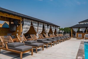 A row of wooden lounge chairs with cushions, a pool, and a covered area with curtains. - The Lighthouse -Steps to Beach!  Luxury Amenities! (Corolla)