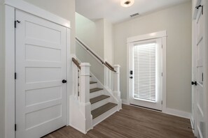 Un couloir avec un escalier en bois, des portes blanches et une fenêtre avec des stores.