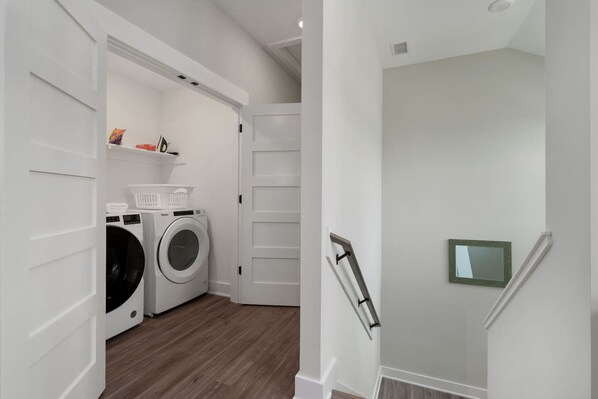 Un espace buanderie comprenant un lave-linge et un sèche-linge, une petite étagère avec des articles de toilette et une porte munie d’une main courante.