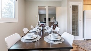 Une salle à manger avec une longue table dressée pour six personnes, des chaises blanches et une vue sur la cuisine.