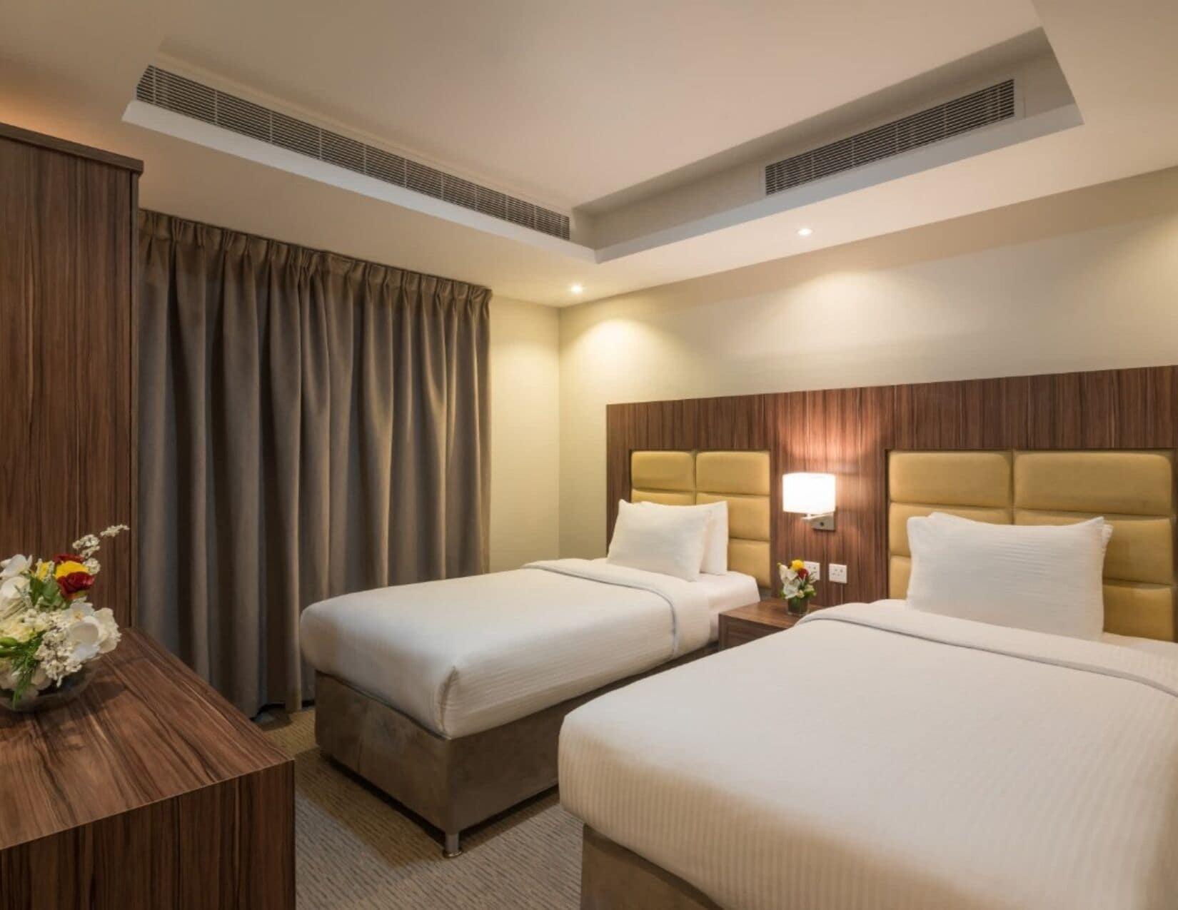 Quarto de hotel com duas camas, uma escrivaninha de madeira, um vaso de flores e uma janela grande com cortinas.