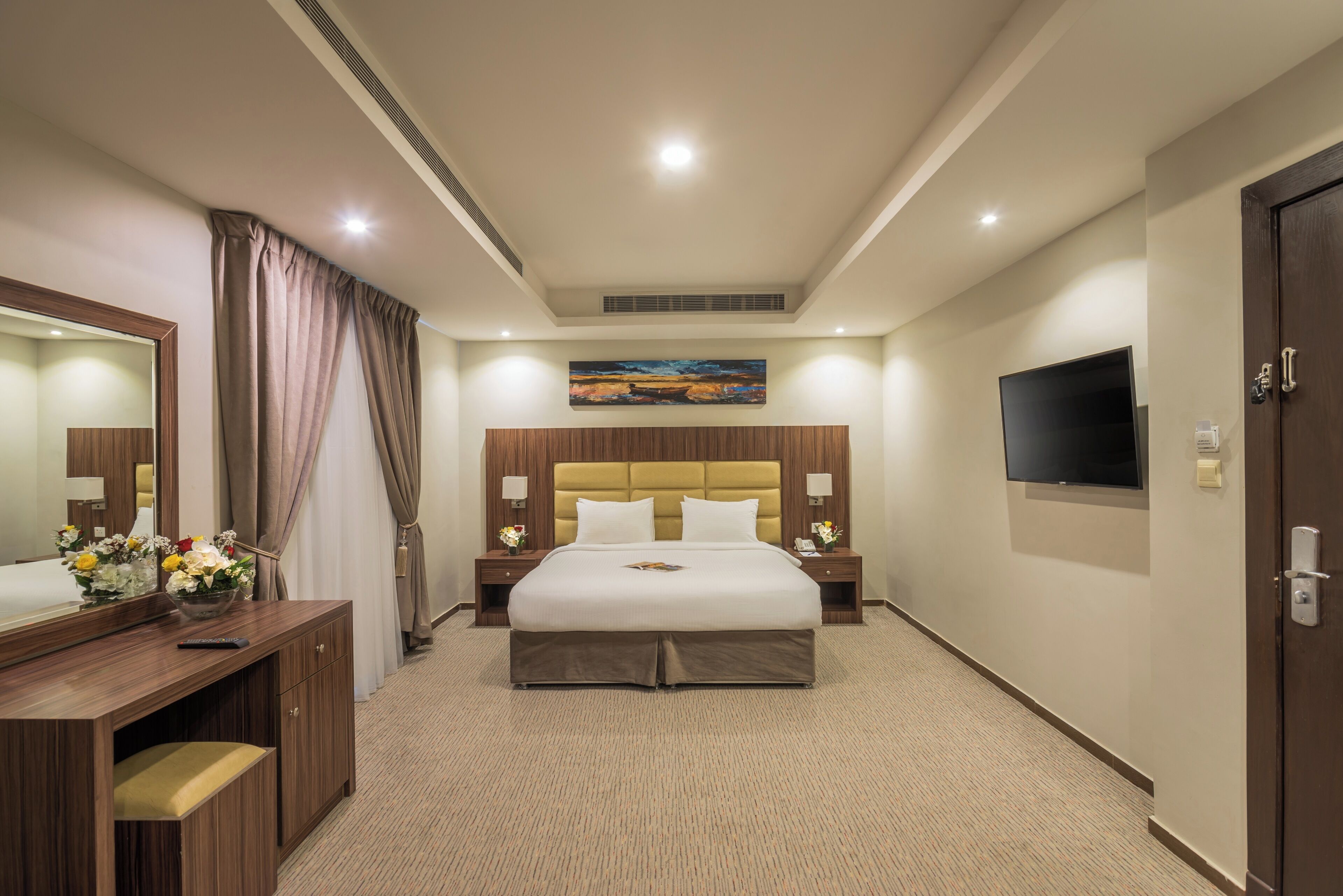 A hotel room with a large bed, a flat-screen TV, a desk with a mirror, and a painting on the wall.