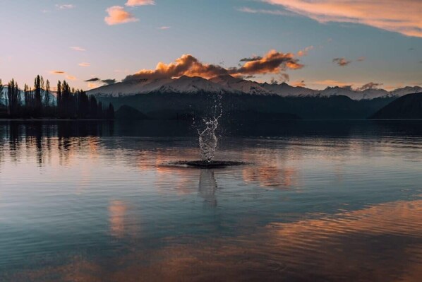 Un lago tranquillo con una fontana al centro, circondato da montagne e alberi.
