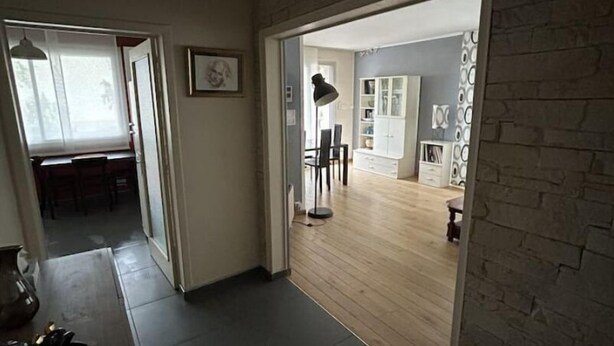 A hallway with a brick wall, a dining area with chairs and a table, and a kitchen area with cabinets and a lamp. - Appartement Cosy (Clermont-Ferrand)
