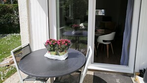 Un patio con una mesa y sillas, un vidrio que da a un espacio interior y un pequeño jardín con flores blancas.