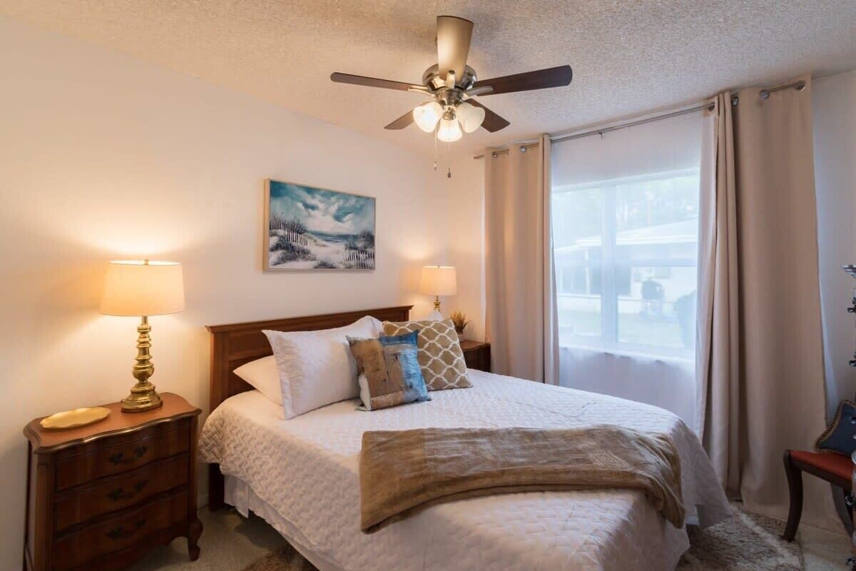 A bedroom with a bed, two bedside lamps, a ceiling fan, a painting on the wall, and a window with curtains.