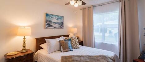A bedroom with a bed, two bedside lamps, a ceiling fan, a painting on the wall, and a window with curtains.