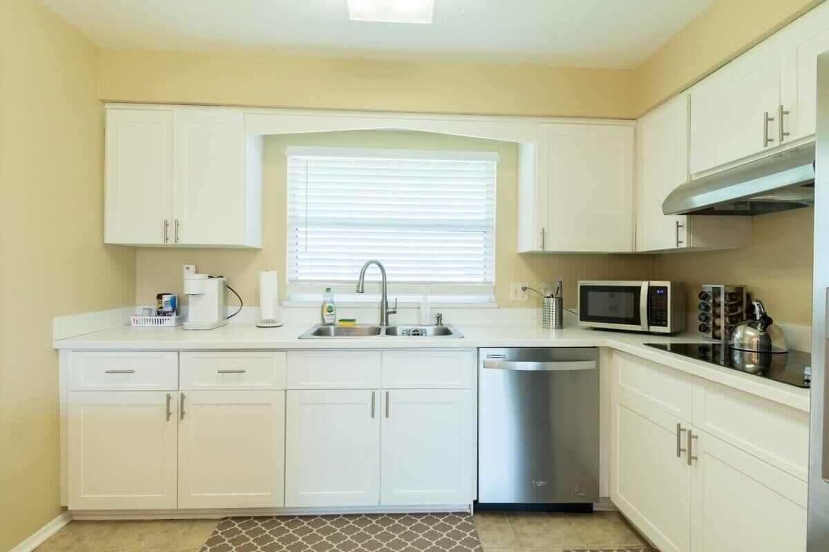 Una cocina funcional con armarios blancos, microondas integrado, lavavajillas y ventana con persianas.
