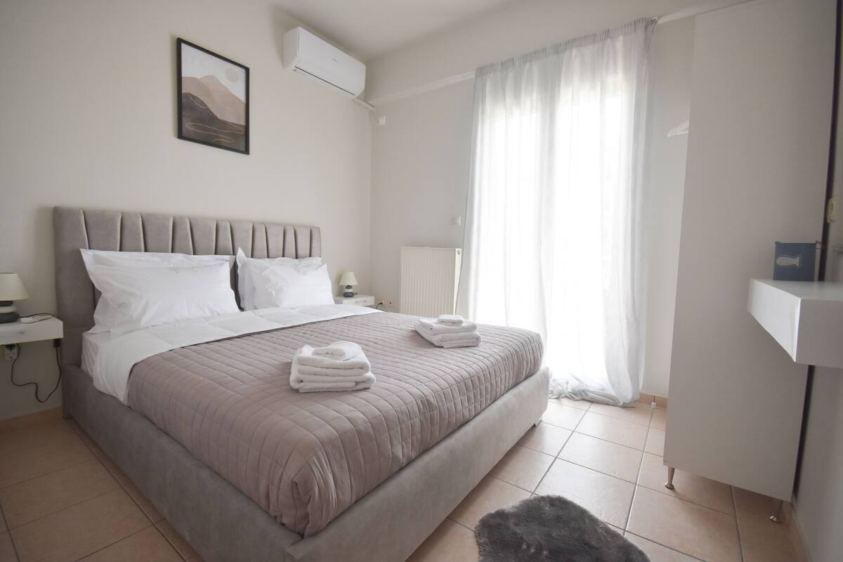A neatly made bed with a beige headboard, white pillows, and folded towels. There is a bedside table with a lamp, a wall-mounted air conditioner, and a framed picture above the bed.