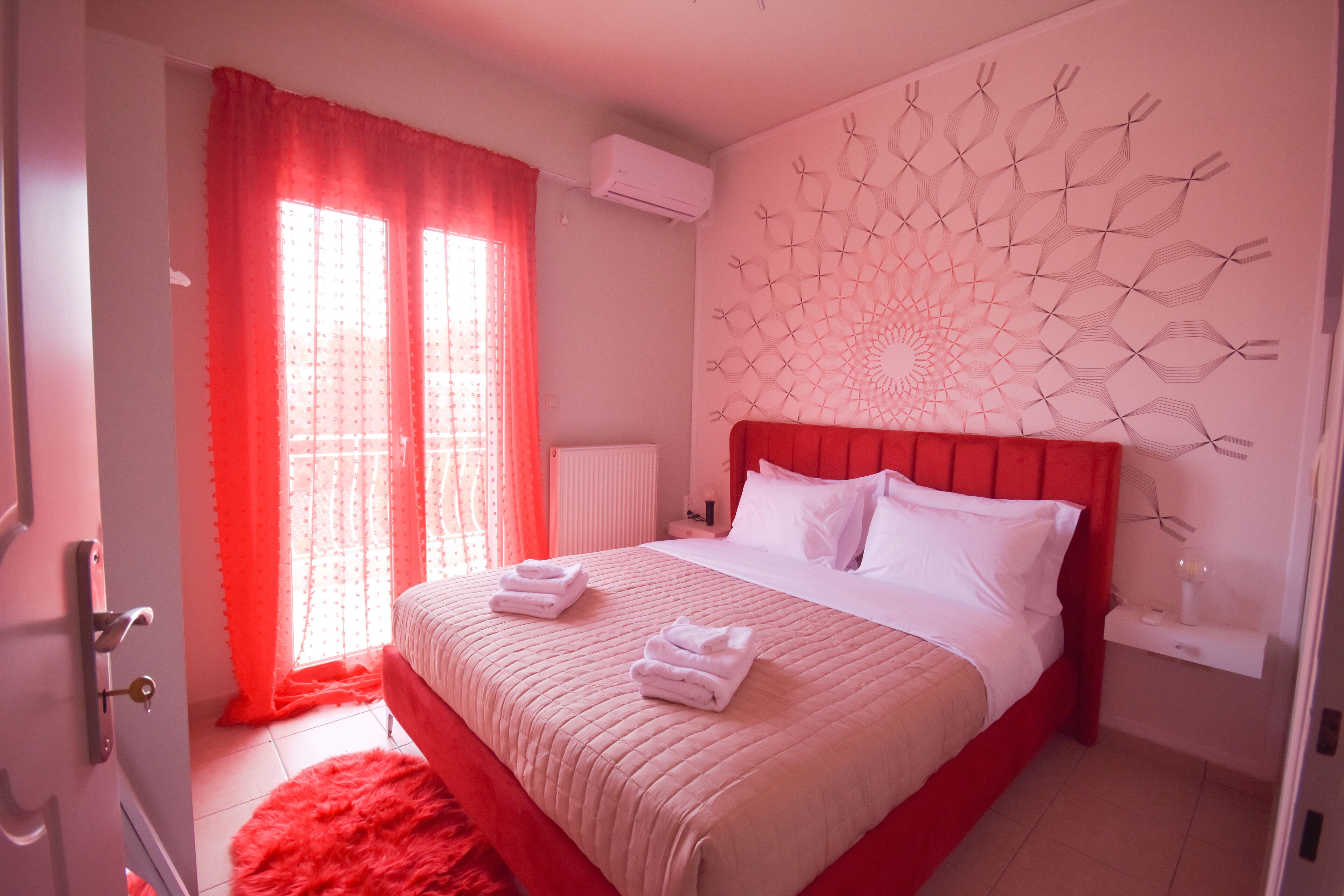 Een hotelkamer met een rood bed, witte kussens en een rood vloerkleed. Er is een raam met rode gordijnen en een decoratie aan de muur boven het bed.