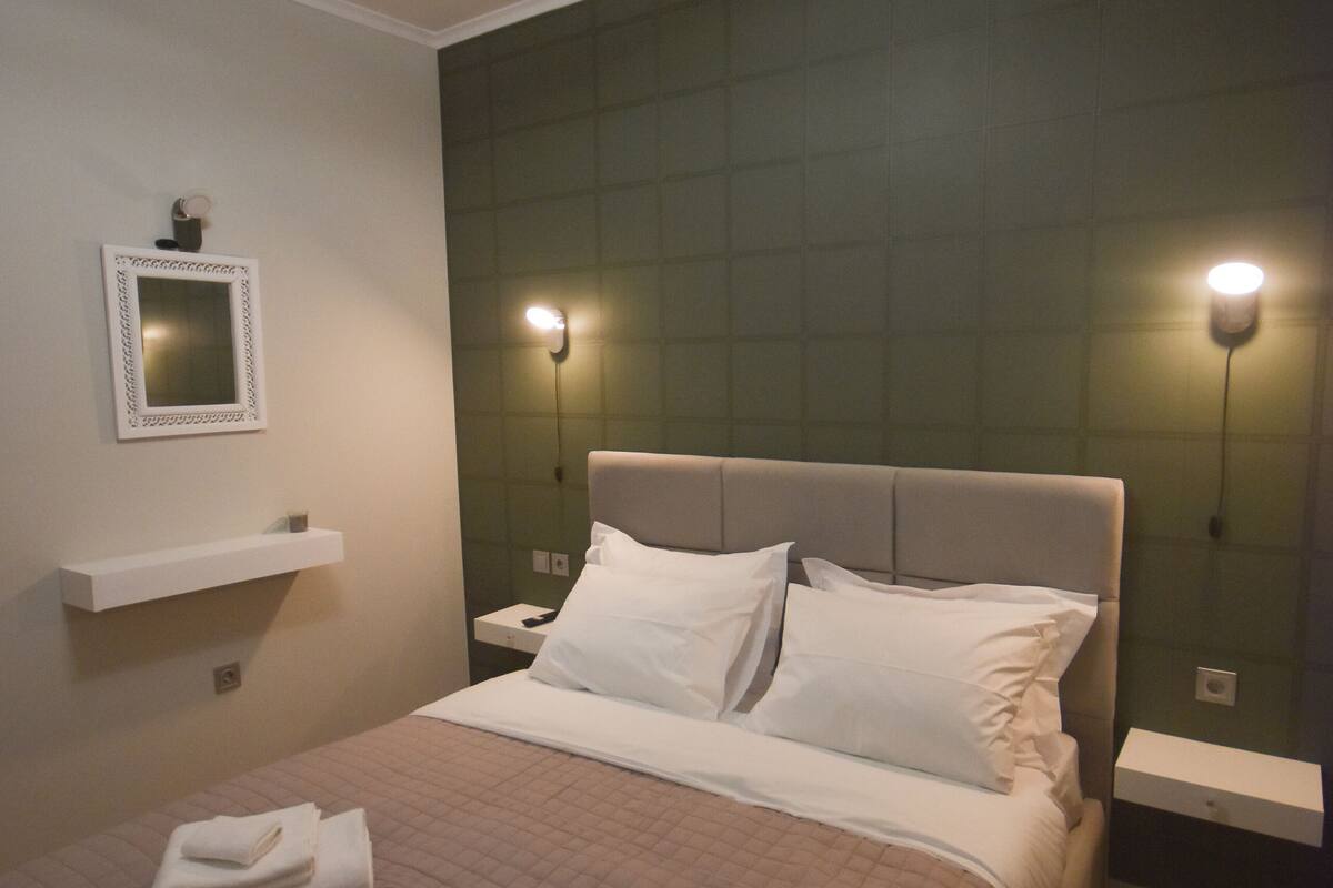 A neatly made bed with a quilted bedspread and two folded towels, a bedside table with a lamp, a wall-mounted shelf, and a mirror.