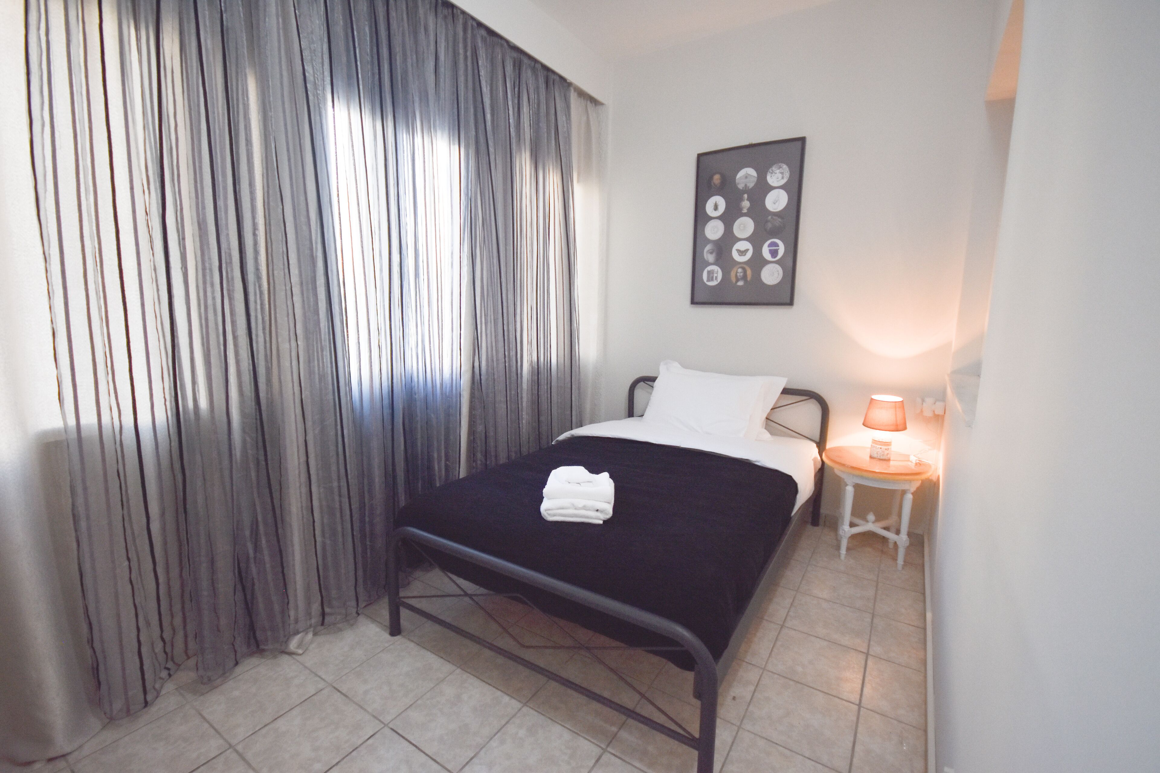 Una cama bien hecha con una colcha negra y almohadas blancas, una mesita de noche con una lámpara y una cortina cubriendo la ventana.