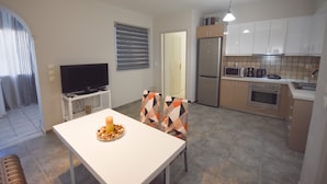 A modern kitchen with white cabinets, a built-in oven, and a microwave. There is a dining area with a white table and chairs, and a television on a stand. - K&C Rooms-Apartments (Markopoulo Mesogaias)