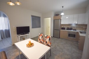 Una cocina moderna con armarios blancos, horno integrado y microondas. Hay un comedor con una mesa y sillas blancas, y un televisor sobre un mueble.