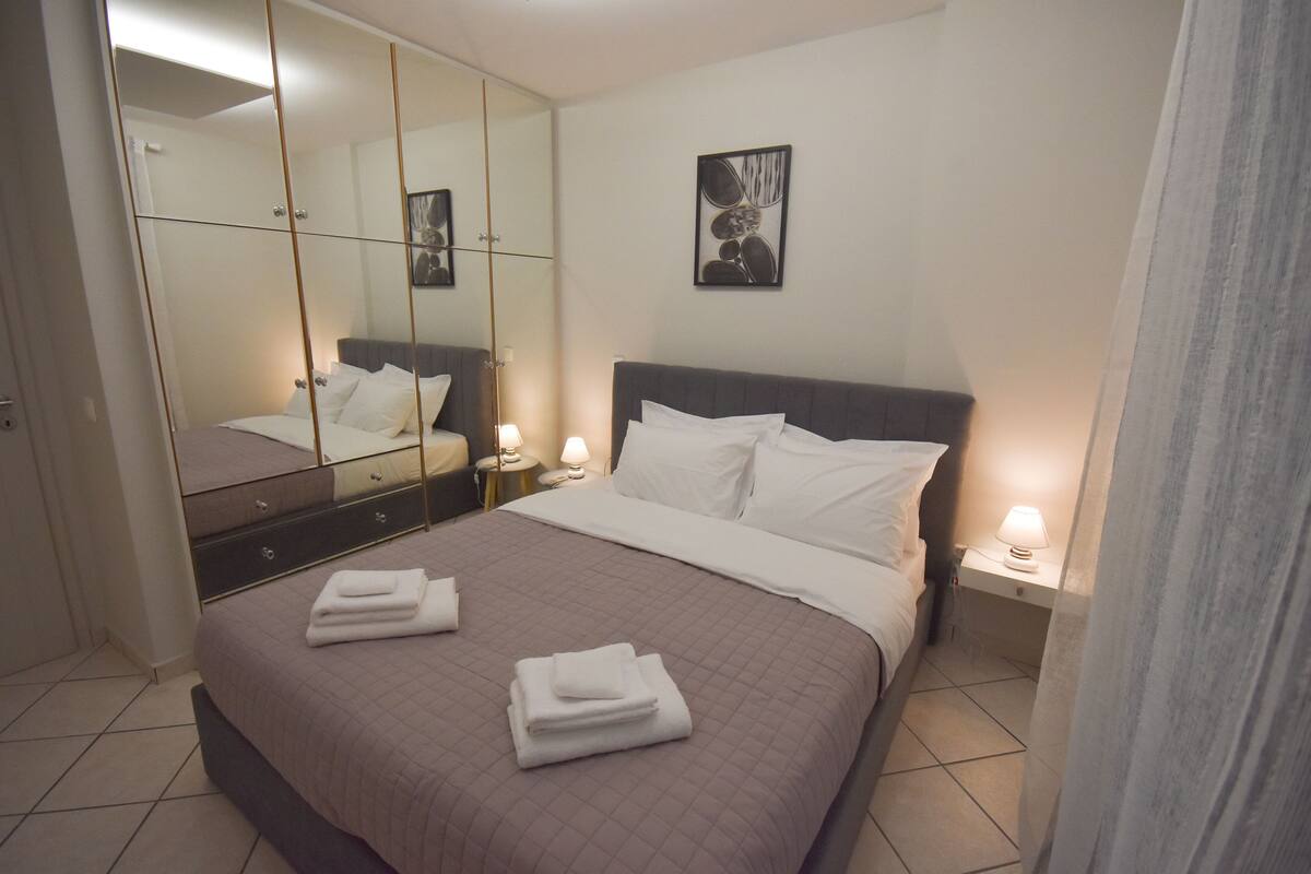 A neatly made bed with white linens and folded towels, a bedside table with a lamp, and a large mirror reflecting the room.