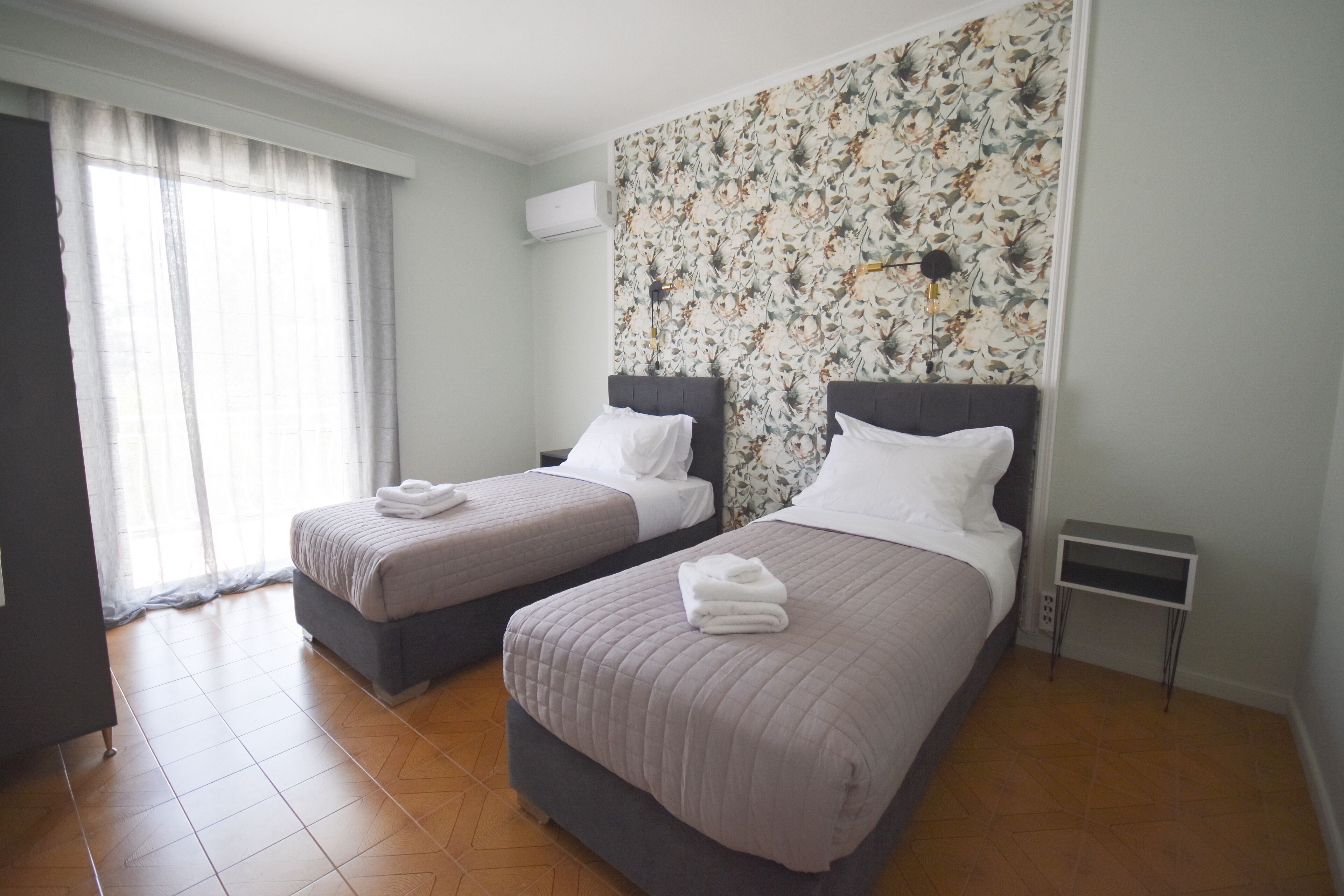 A hotel room with two beds, a floral wallpaper, a small bedside table, and a window with curtains.