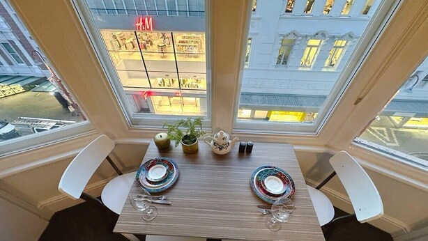 A dining area with a table set for two, a view of a street with a store named 'H&M,' and a building with 'BRADES' on it. - Central Collins Residence at Inner City (Hobart)