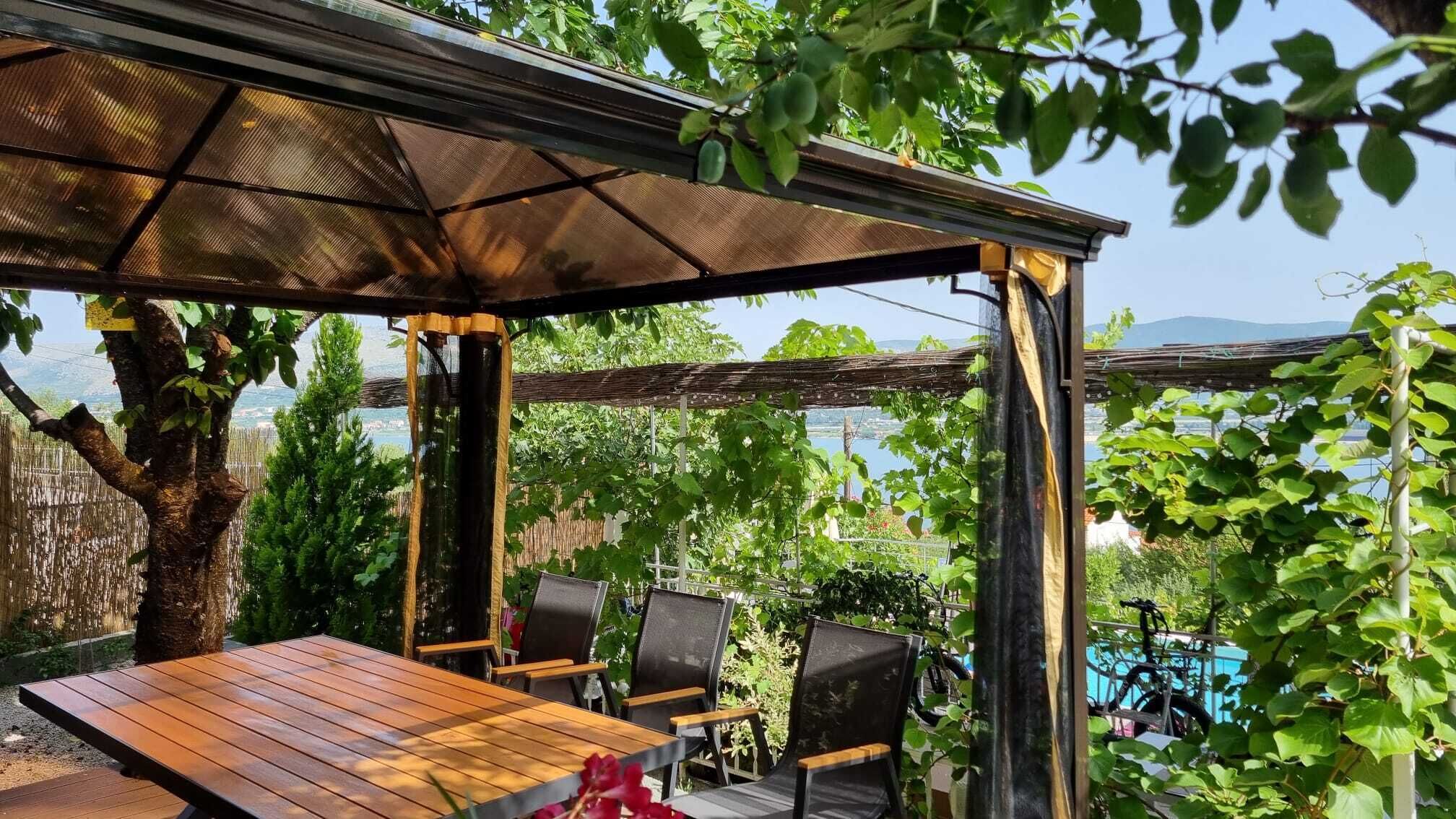 An outdoor seating area with a pergola, wooden table, and chairs, overlooking a garden and a body of water.