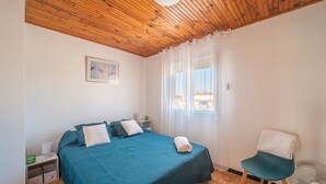A bedroom with a wooden ceiling, a bed with teal bedding, a chair, a bedside table with a lamp, and a window with curtains. - Une Maison à la Mer - Proche de la marina (Saint-Cyprien)