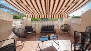 An outdoor patio with a striped canopy, black metal chairs, a table with a cup, and a view of greenery in the background. - Une Maison à la Mer - Proche de la marina (Saint-Cyprien)