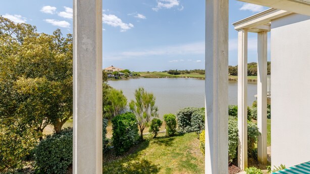 A view from a covered porch overlooking a lake, with a white building and greenery in the foreground. - L'Hollywood Zen - One Bedroom Apartment, Sleeps 4 (Saint-Jean-de-Monts)