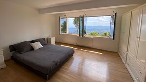 Une chambre à coucher comprenant un lit, une table de chevet, une armoire et de grandes fenêtres donnant sur l’extérieur.