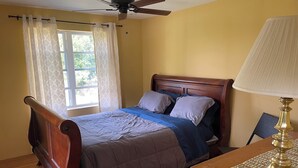 A bedroom with a wooden bed, a ceiling fan, a window with curtains, a bedside table with a lamp, and a chair.