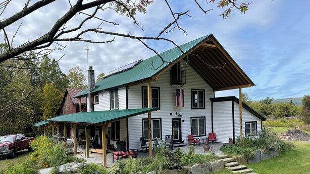 A two-story house with a green roof, a porch with red chairs, and a stone pathway leading to the front door.