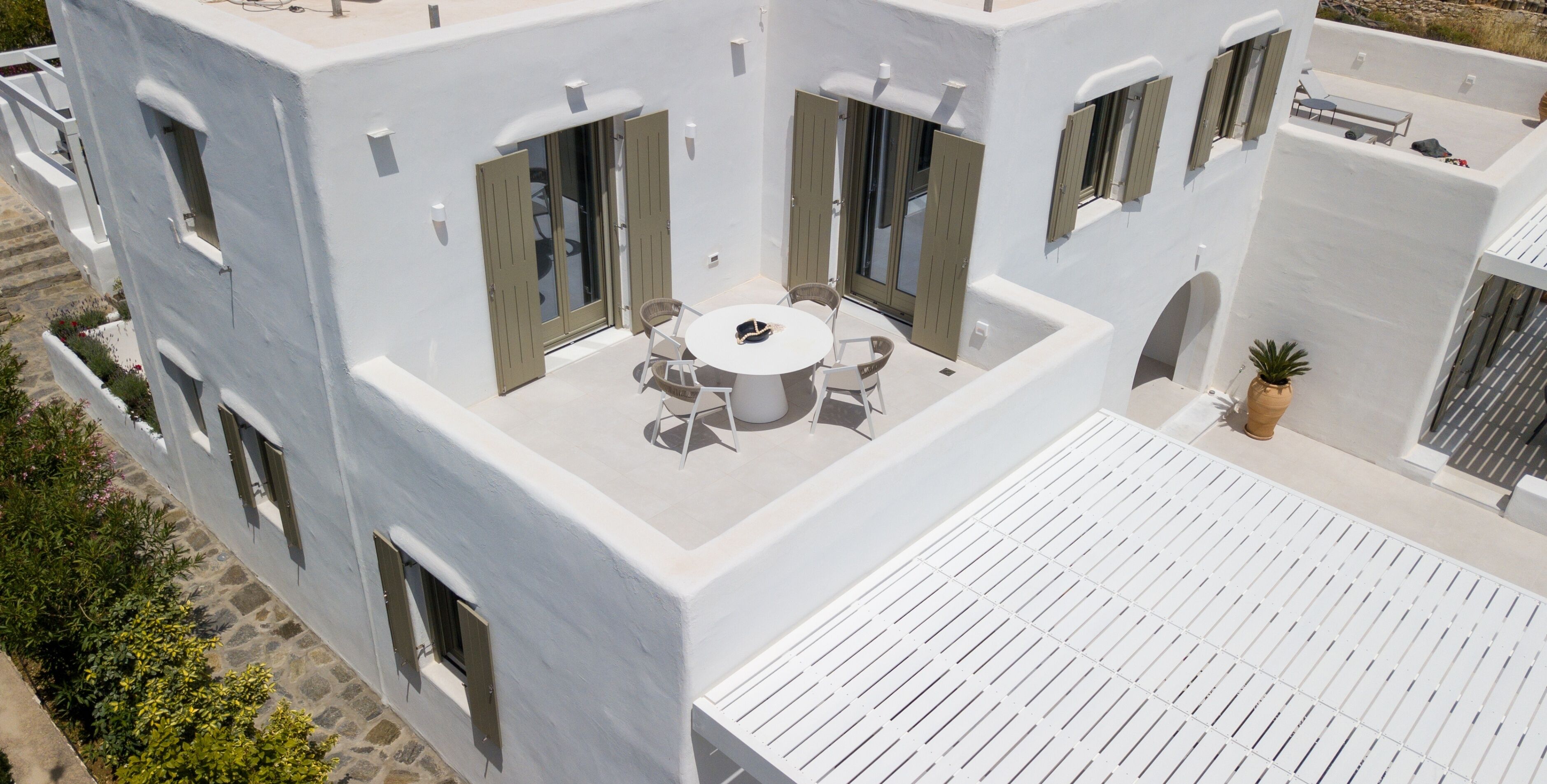 A modern building with a rooftop terrace featuring a round table and chairs, a potted plant, and a white railing.