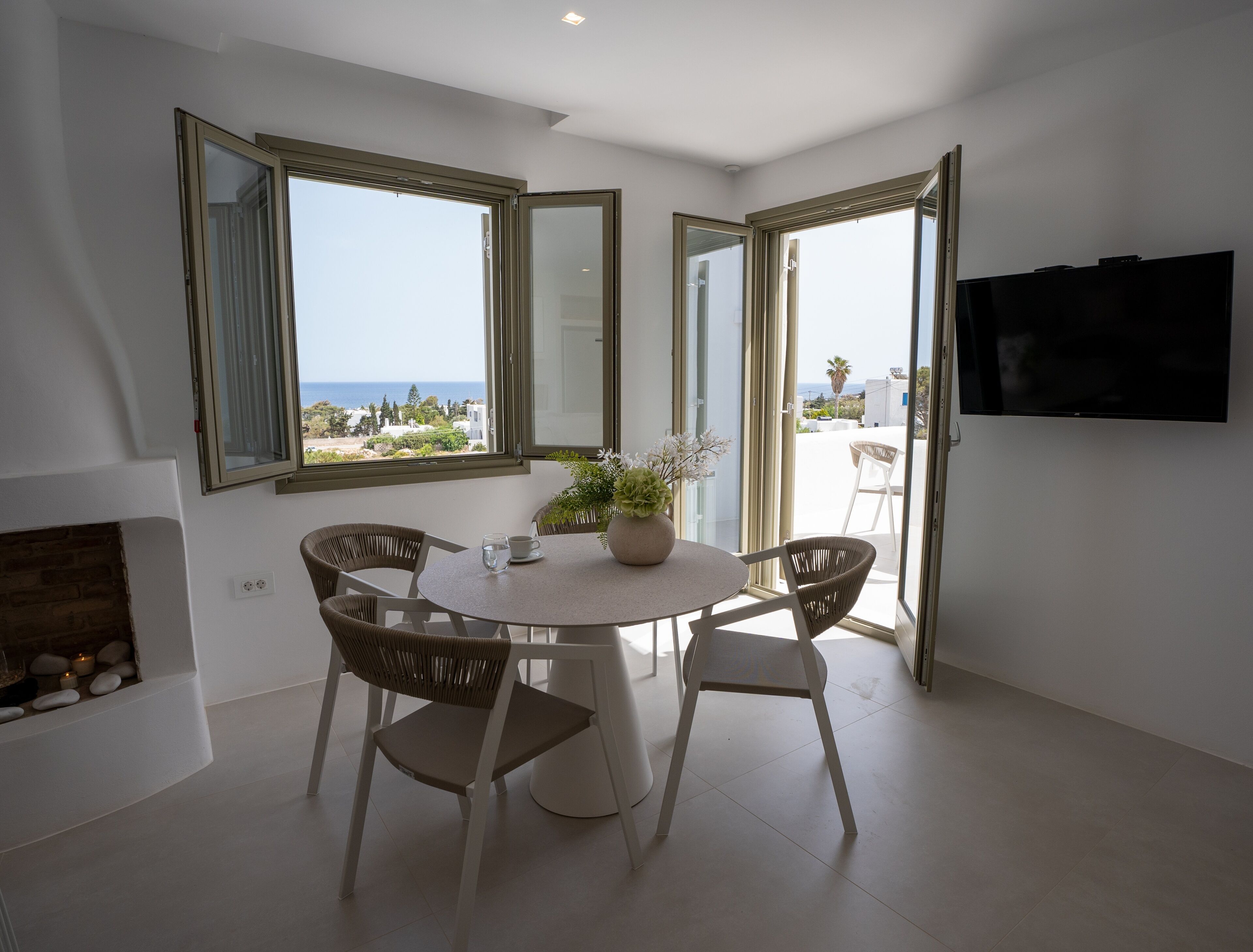 A modern living room with a round table, wicker chairs, a TV, and large windows offering a view of the sea.