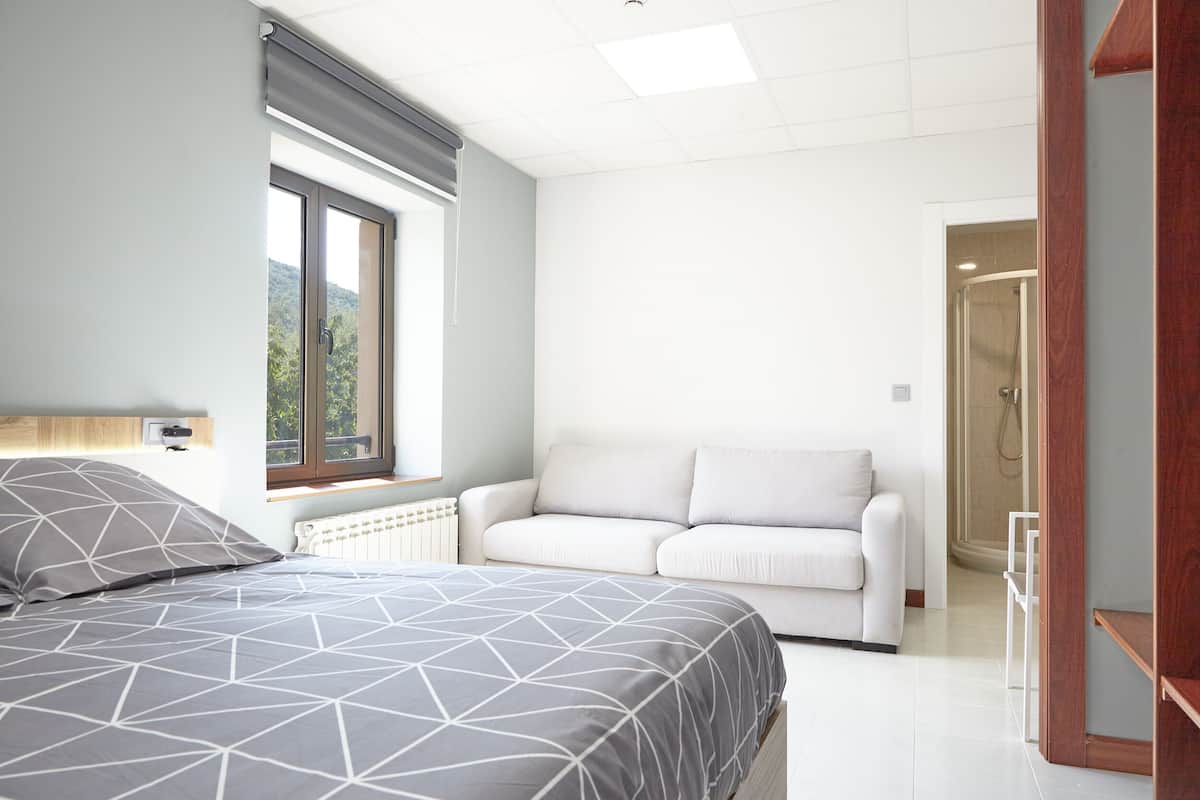 Un dormitorio con una cama, un sofá blanco, un radiador y una ventana con vistas a zonas verdes.